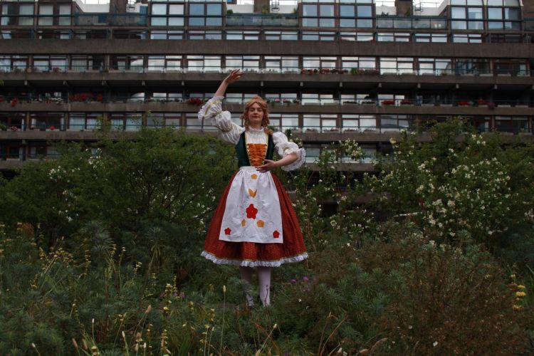 Model posing in a ballet position with a traditional style female dress - white shirt with puffy sleeves, dark green bodice with red corset style tie criss-crossing the front, red skirt and white apron embroidered with red and orange flowers
